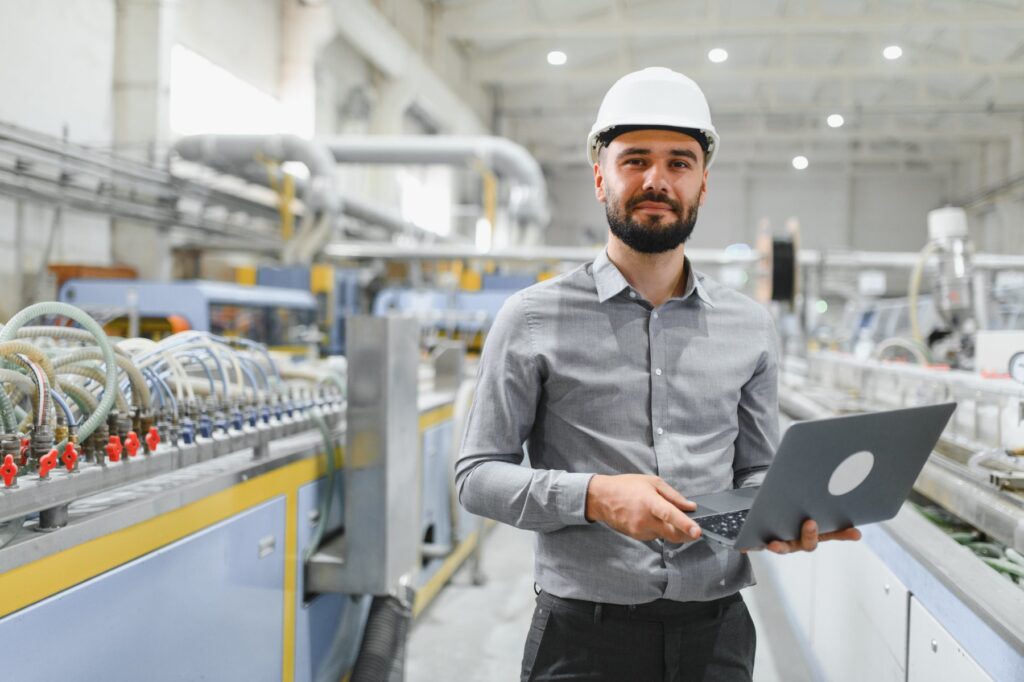 Manufacturing engineer in a hard hat holding a laptop while overseeing a centralized production line